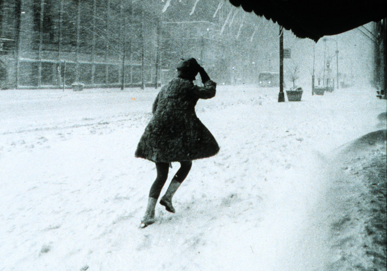 image of a woman in a green dress running away from the viewer in a snowstorm. she is partially under a dark awning, behind her, her footprints make tracks in the new snow.