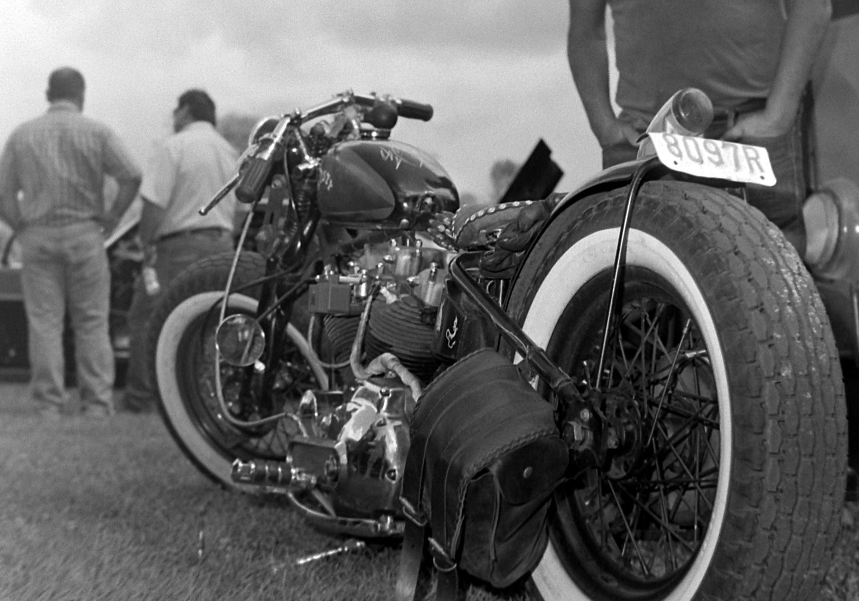 close up image of an old motorcycle propped up in grass, with a man with his hands in his pockets standing behind it, and two men far off in the left hand corner of the frame looking away.